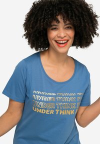 Camiseta corta de manga azul, hecha de una tela suave, que presenta un atrevido texto dorado en múltiples tipografías: "UNDER THINK IT" en el pecho, en patrones repetidos.