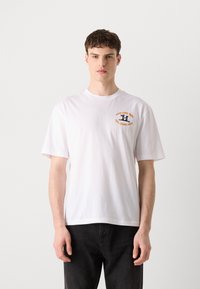 T-shirt bianco in cotone con maniche corte, caratterizzato da una stampa grafica arancione sul lato sinistro del petto. Orlo dritto. Modello che indossa pantaloni neri.
