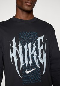 Schwarzes Langarmshirt mit einem großen weißen und blauen Nike-Logo, das ein Netzdesign im Hintergrund und silberne Akzente aufweist.