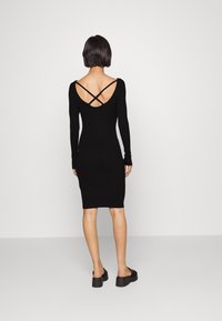 Musta pitkähihainen bodycon-mekko, jossa on syvä V-muoto takana ja ristikkäiset olkaimet. Mekko on vartalonmyötäinen ja pituus ylettyy pohkeisiin.
