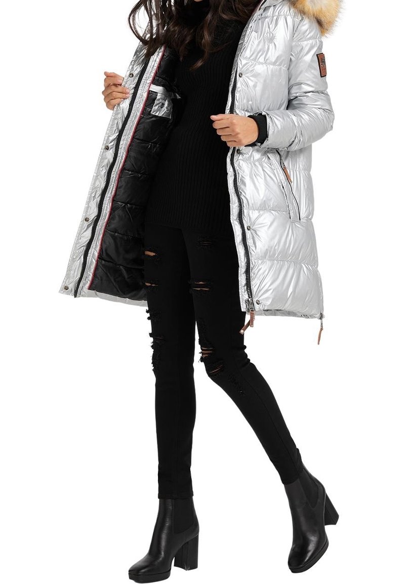 Femme portant une doudoune argentée brillante avec capuche bordée de fourrure, un jean skinny noir déchiré, un pull noir et des bottines à talons noires.