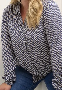Femme portant un chemisier à manches longues bleu et beige à motif avec des boutons, et un jean bleu, assise avec une main reposant sur le genou.