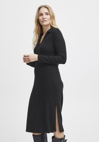 Svart ribbad långärmad klänning med V-hals och sidoklyftor, gjord av mjukt tyg, figurformad silhuett och knälång design.