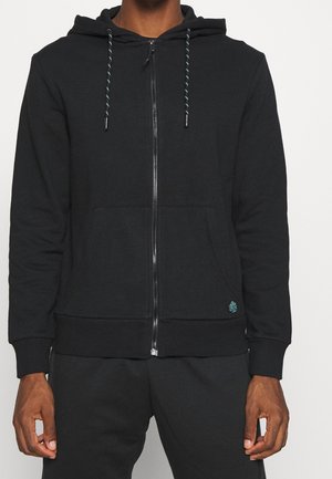 Mężczyzna ubrany w czarną bluzę z kapturem na suwak, z sznurkami i kieszeniami z przodu, stojący na jasnym, gładkim tle.