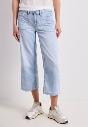 Licht gewassen cropped wijde jeans, gedragen met witte en zilveren sneakers en een wit topje, weergegeven vanaf halverwege het bovenbeen naar beneden tegen een effen achtergrond.