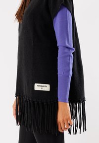 Poncho in misto lana nera con orlo frangiato, caratterizzato da un'etichetta con logo. Indossato sopra una maglietta a maniche lunghe viola, mostrando colori e trame contrastanti.