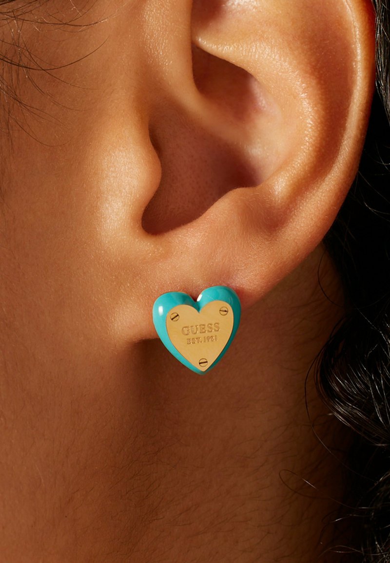 Türkisfarbene, herzförmige Ohrringe mit goldenen Akzenten, graviert mit "GUESS EST. 1981", sicher am Ohrläppchen befestigt. Glatte Oberfläche.