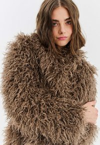 Mladá žena se středně dlouhými hnědými vlasy, oblečená v tlustém, kudrnatém hnědém kožichu, hledící přímo do kamery s neutrálním výrazem.
