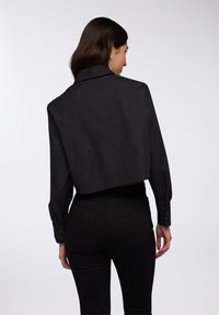 Maglietta nera a maniche lunghe, cropped, con polsini abbottonati e un collo ampio. Tessuto liscio e vestibilità su misura. Abbinata a jeans neri a vita alta.