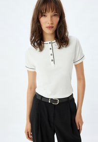 Camisa polo blanca de manga corta, con ribete negro y tapeta de cuatro botones. Combinada con pantalones negros de cintura alta y un cinturón negro delgado.