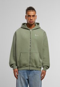 Olive green zip-up hoodie in soft fabric, featuring a kangaroo pocket, ribbed cuffs, and a subtle logo on the left chest.