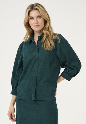Camicia di velluto a coste color turchese con chiusura a bottoni, colletto e maniche a sbuffo. Presenta bottoni marroni e una finitura testurizzata.