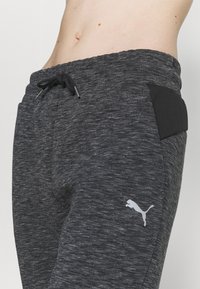 Zbliżenie na talię osoby noszącej ciemnoszare spodnie dresowe Puma z sznurkiem i logo na lewej udzie.