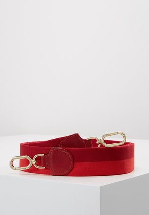 Sangle large en tissu rouge avec fermoirs métalliques dorés aux deux extrémités, avec renfort en cuir rouge près de chaque fermoir, enroulée sur une surface blanche.