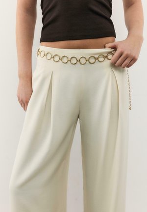 Person wearing cream wide-leg pants with pleats, a gold chain belt with circular links, and a black cropped top.
