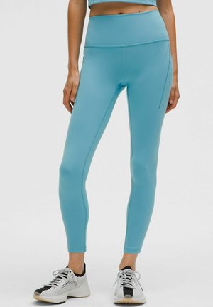 Een persoon in een aansluitend blauw top en bijpassende leggings staat, en toont athletic schoenen en een stijlvolle, sportieve uitstraling.