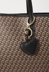 Tote bag with a brown and beige geometric pattern, black leather accents, and a black heart-shaped charm attached by gold hardware.