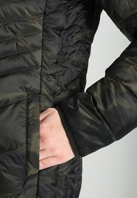 Veste rembourrée présentant un motif camouflage dans des nuances de noir et de vert foncé, avec des sections matelassées et des poches latérales.