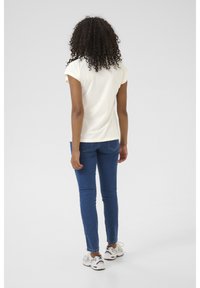 Kortærmet creme t-shirt, tætsiddende design, parret med blå skinny jeans og hvide sportssko, set fra ryggen.