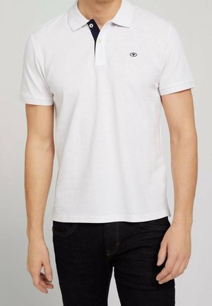 Mężczyzna ubrany w biały polo z krótkim rękawem, z małym logo na lewej piersi, w połączeniu z czarnymi spodniami.