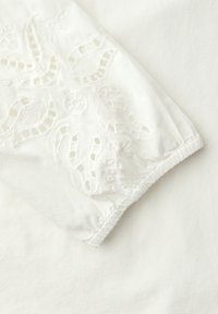Manche en tissu blanc avec un poignet élastique ornée de broderies ajourées florales et de coutures texturées sur un fond blanc uni.
