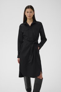 Hosszú, sötét hajú nő fekete hosszú ujjú ruhát visel derékkötővel és fekete térdig érő csizmával, egyszerű háttér előtt állva.