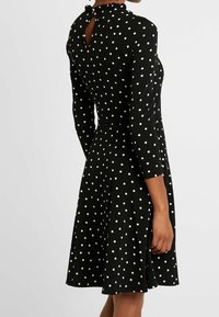 Musta mekko, jossa pitkät hihat, valkoiset pilkut, vartalonmyötäinen yläosa, levenevä helma ja röyhelöinen pääntie, jossa avainreikäleikkaus takana.