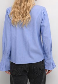 Haut à manches longues bleu clair avec des rayures blanches verticales, featuring des manches ballons et des poignets évasés, associé à un jean gris foncé.