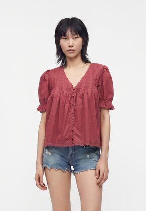Jeune femme aux cheveux noirs portant un chemisier rouge à manches courtes avec des boutons et un short en jean bleu délavé, sur fond uni.