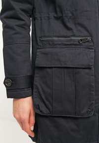 Main reposant à côté d'une manche de veste noire avec un poignet boutonné, montrant une poche zippée au-dessus d'une grande poche à rabat à l'avant de la veste.
