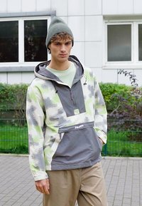 Casaco leve tipo pulôver com um padrão de camuflagem em verde, cinza e branco. Apresenta um bolso frontal, capuz com cordão e um detalhe com logo.