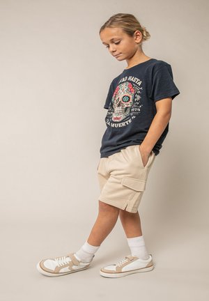 Kind met handen in zakken, draagt een zwart t-shirt met schedelprint, beige cargoshorts, witte sokken en witte sneakers, staand tegen een beige achtergrond.
