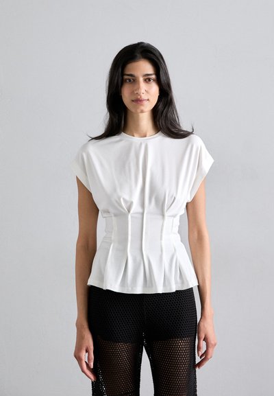 Top peplum alb cu mâneci scurte și detalii pliate, asortat cu colanți din plasă negri, cu un model deschis de tricot.