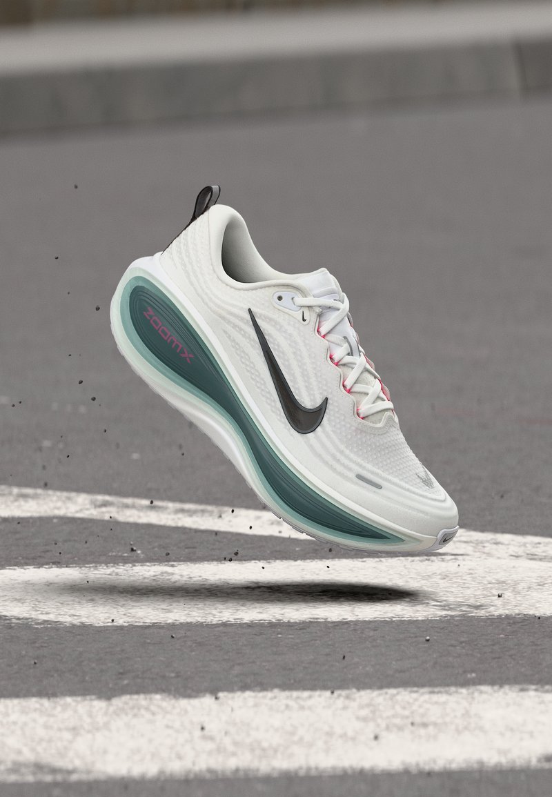 Λευκό αθλητικό παπούτσι Nike ZoomX με μαύρο λογότυπο, αιωρούμενο πάνω από άσφαλτο με λευκές σημάνσεις δρόμου και διάσπαρτα μικρά συντρίμμια.