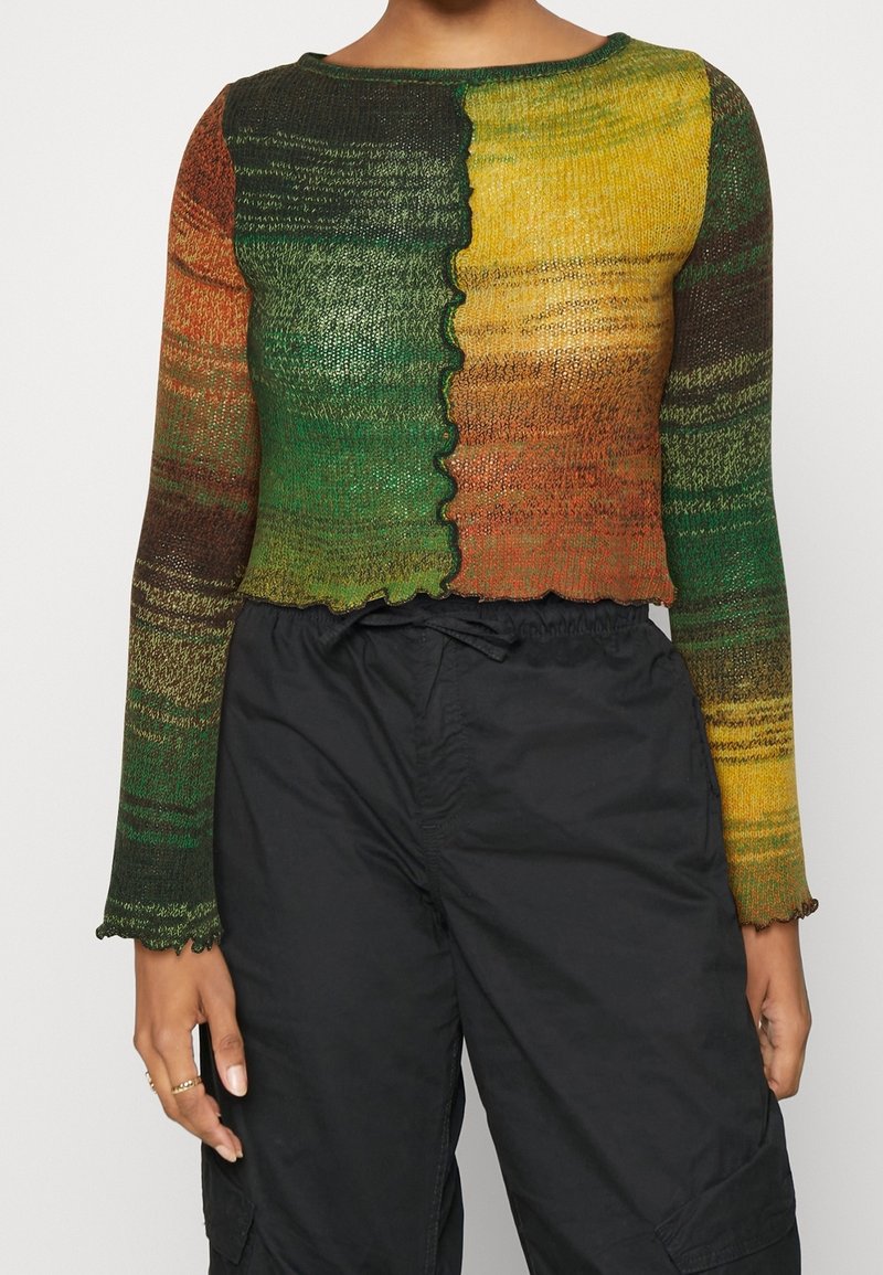 Haut en maille multicolore à manches longues vert, jaune et orange avec couture frontale ondulée et bords volantés, associé à un pantalon noir à taille élastique.