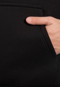 Sudadera negra con un bolsillo tipo canguro, tela texturizada y detalles de costura visibles, con una mano colocada en el borde del bolsillo.
