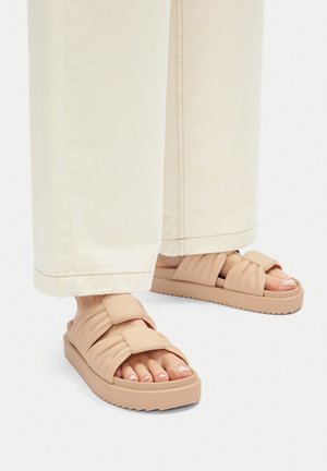 Pies con sandalias tipo slide acolchadas beige con tiras anchas y suelas gruesas, combinadas con pantalones anchos de color crema.