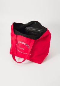 Sac fourre-tout en toile rouge avec intérieur noir, deux poignées, poche zippée et marquage blanc sur le devant, présentant un design minimaliste.
