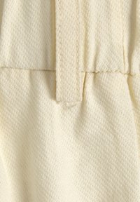 Tessuto di crema testurizzato con un aspetto morbido e intrecciato. Presenta arricciature e un legaccio centrale, che enfatizza una finitura sartoriale.