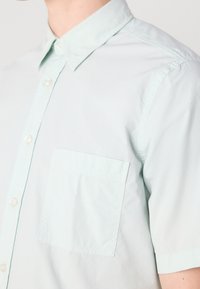 Hombre vistiendo una camisa de manga corta color menta pálido con botones blancos, bolsillo en el pecho y cuello.