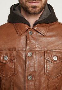 Bruine leren jas met een gestructureerde afwerking, zilveren drukknoopjes en twee borstzakken, gelaagd over een donkergrijze hoodie.