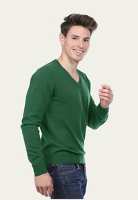 Pull vert en v, en maille douce et texturée, avec des manches longues et une coupe ajustée. Porté avec un jean en denim foncé.