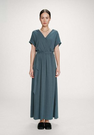 Femme debout, portant une robe longue style portefeuille bleu sarcelle à manches courtes, une ceinture à la taille, des chaussures noires sans lacets, et un collier chaîne en or.