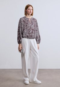 Μπλούζα με λεοπάρ σχέδιο, στρογγυλή λαιμόκοψη και λεπτομέρεια με άνοιγμα, σε συνδυασμό με αέρινο λευκό παντελόνι και λευκά αθλητικά παπούτσια, κρατώντας μια μικρή ασημένια τσάντα.