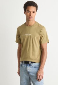 Olive green cotton t-shirt with short sleeves, round neckline, and white text logo in the center. Paired with light blue denim jeans.