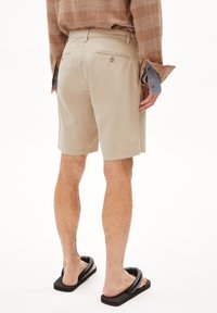 Shorts beiges ajustés avec poches arrière, fermeture à bouton, jambes droites. Portés avec des sandales noires à enfiler, présentant une chemise texturée couleur caramel.
