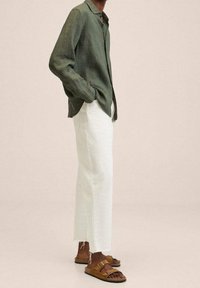 Leinenhemd in Olivgrün, locker geschnitten; weite weiße Leinenhose mit ausgefranstem Saum; braune Sandalen mit zwei verstellbaren Riemen.