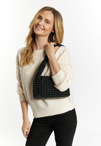 Schwarze gewebte Tasche mit einzigartigem strukturiertem Design, dicken Trägern und dreieckiger Form, gehalten von einer Person, die einen hellen Pullover und dunkle Hosen trägt.