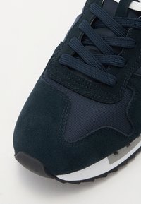 Zapatillas de color azul marino que combinan materiales de ante y malla, con cordones planos, una suela texturizada y detalles en blanco y gris a contraste.