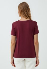 Donna con capelli biondi lisci che indossa una semplice maglietta rossa scura a maniche corte e pantaloni bianchi, vista di schiena su uno sfondo bianco.